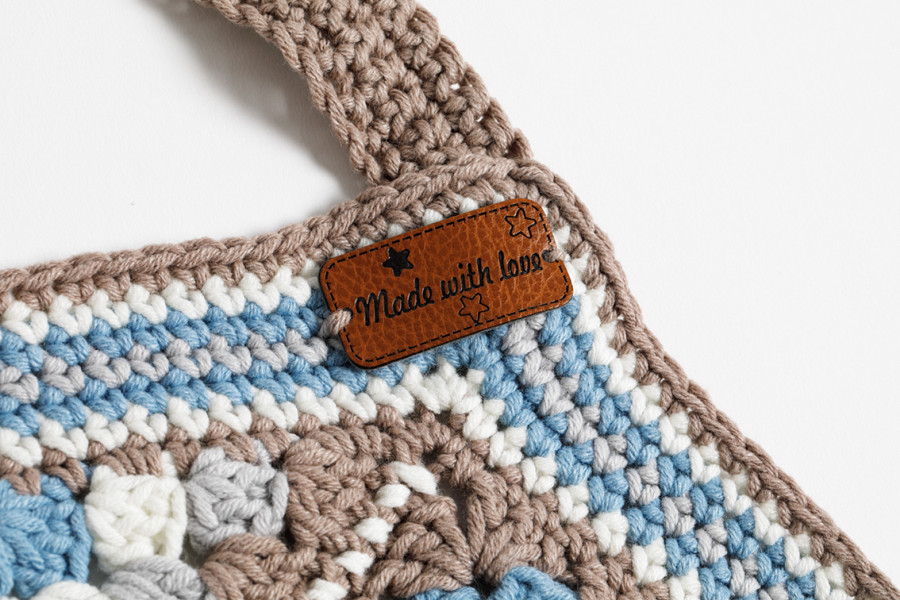Bovenkant van het gehaakte schort van granny squares. De kleuren taupe, ivoor, lichtblauw en grijs worden gebruikt. In het rechterbovenhoekje van het schort is een bruinleren label bevestigd waarop staat: 'Made with love'.